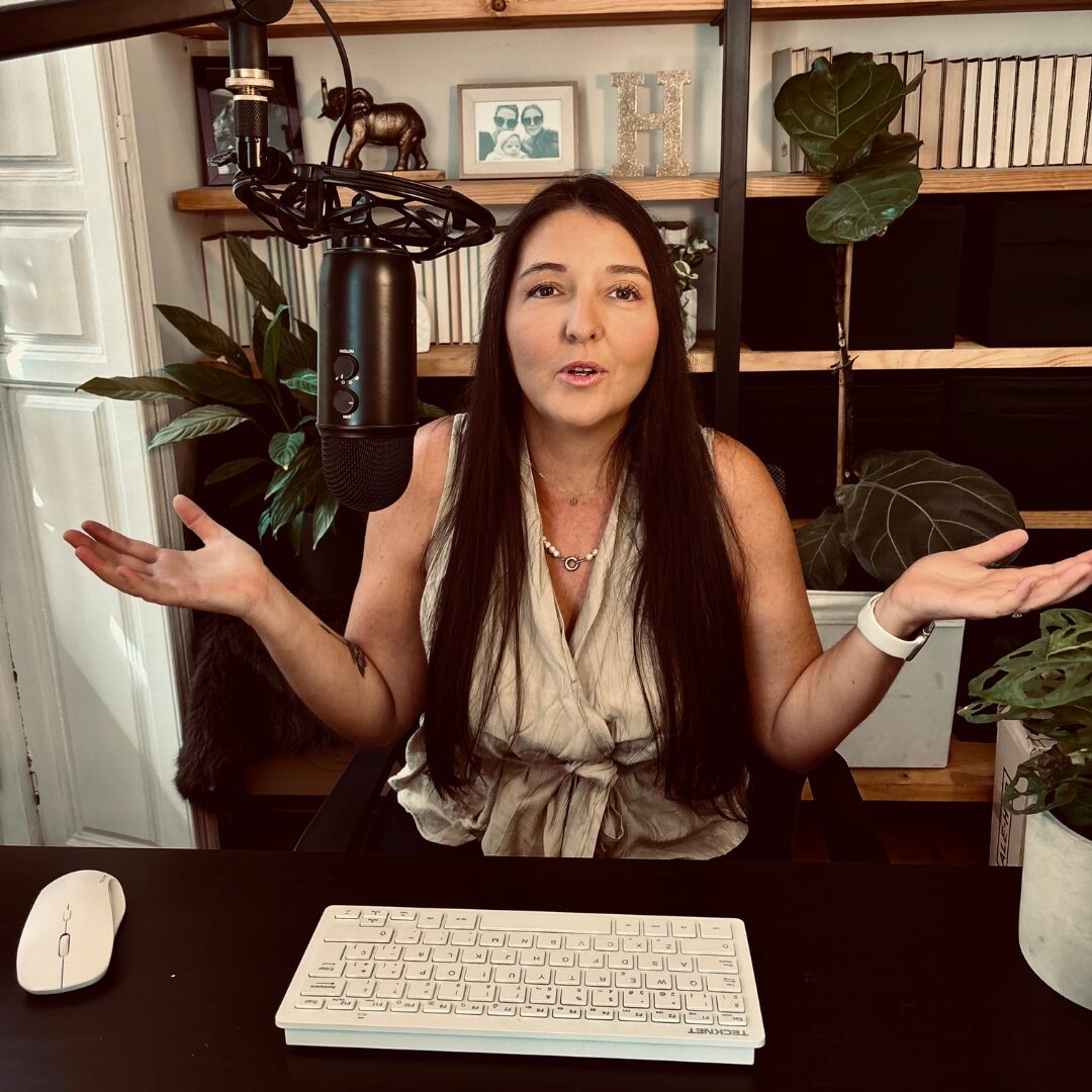 Heidi Hein Pickering, site editor of Expat Wife Hustle, seated at her desk in front of a microphone, appearing to ask the question "how we make money?" Her workspace includes a laptop and microphone, indicating she’s recording or working on content creation."